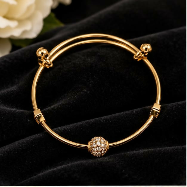 A gold-tone adjustable bangle bracelet with a sparkling crystal-studded ball charm centered on a black velvet surface.