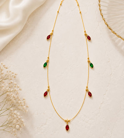 The gold-plated chain necklace with red and green crystal pendants is laid flat on a light textured surface, accompanied by