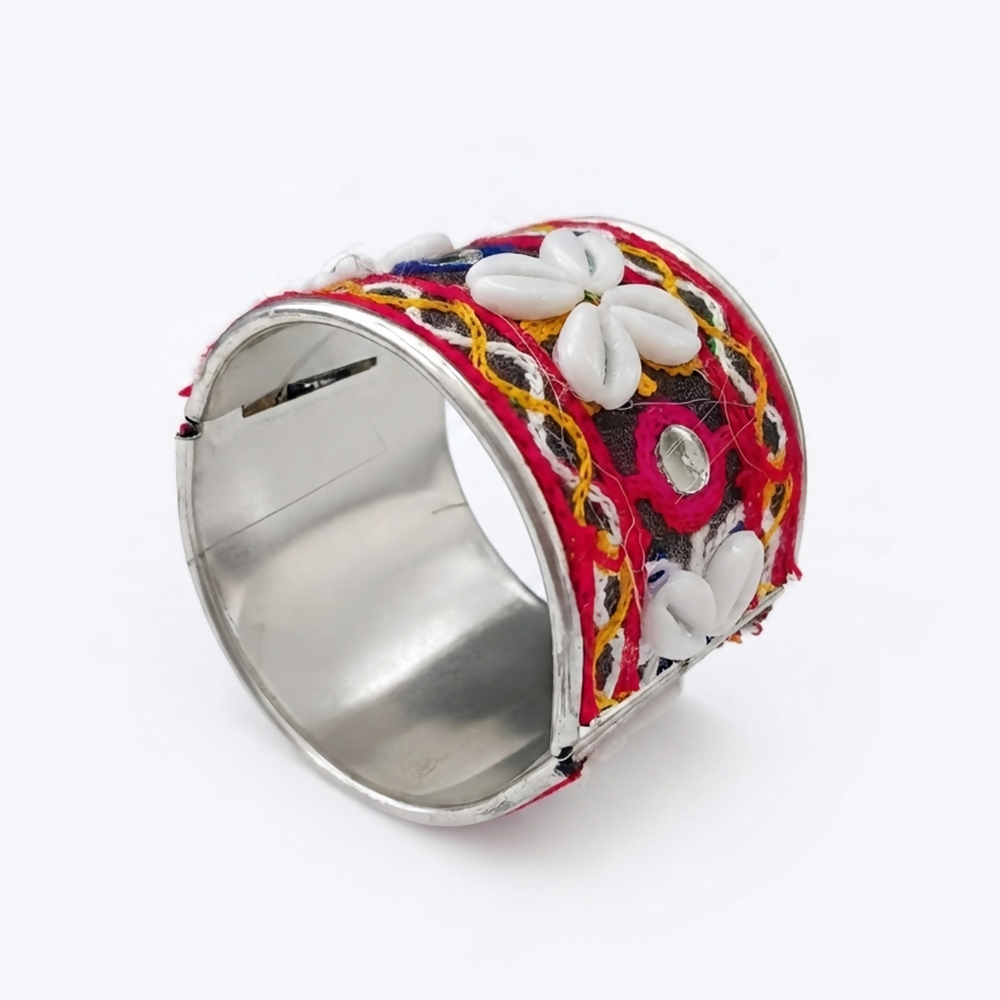An open silver cuff bracelet displays intricate red, yellow, and blue embroidery, reflective mirrors, and white cowrie shell