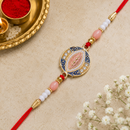 A blue, pink, and gold Rakhi with a central oval design, surrounded by small white stones and pearls, rests on a light brown