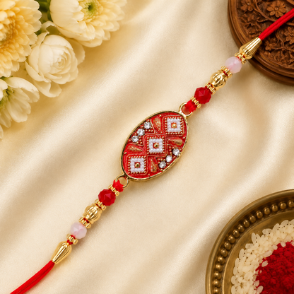 A red and gold designer Rakhi with a central oval charm featuring red, white, and gold patterns, accented with clear stones.