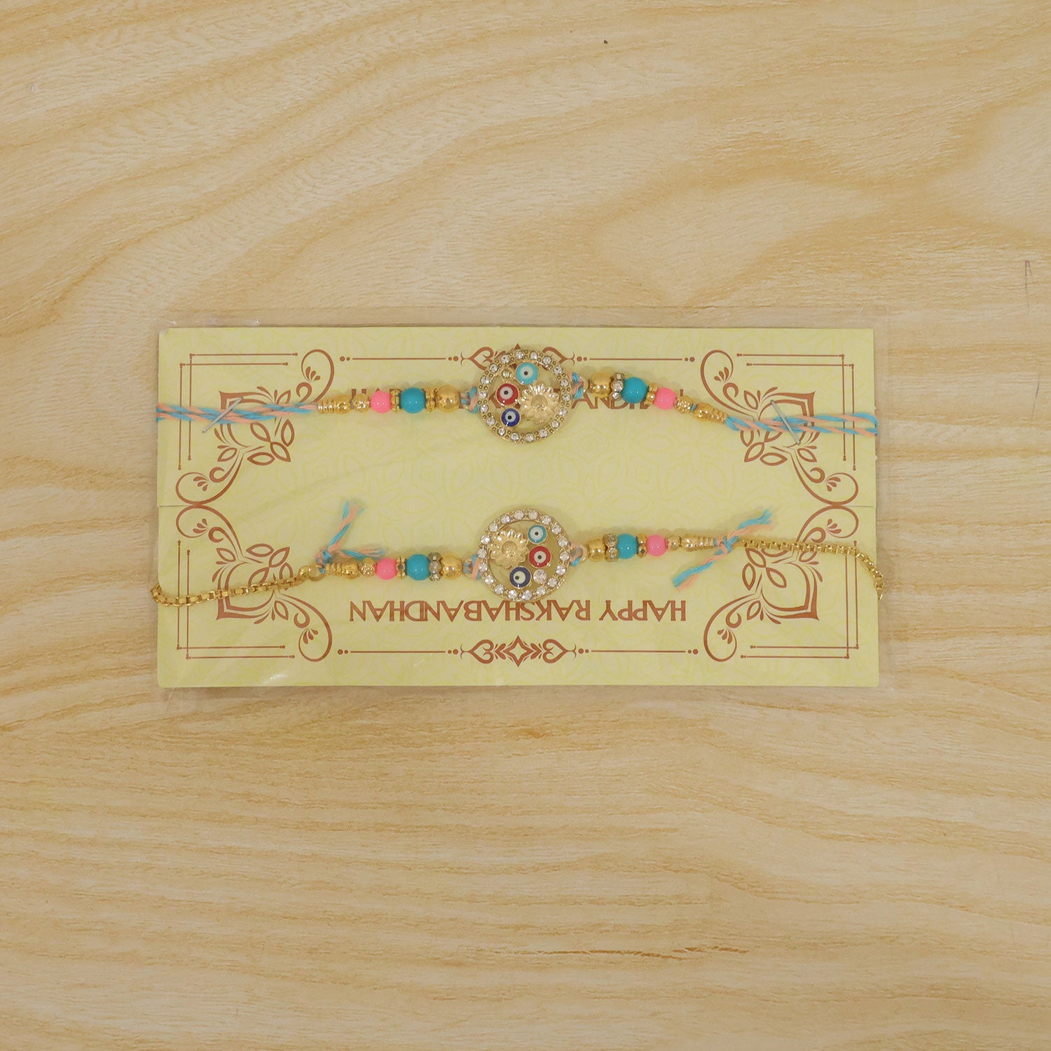 Two colorful Rakhi bracelets with evil eye beads and a gold lumba, packaged on a yellow card with text, on wood.