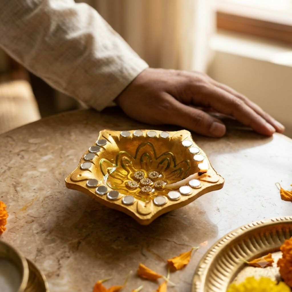 A gold diya with mirror accents and a floral design rests on a light marble surface next to a person's arm.