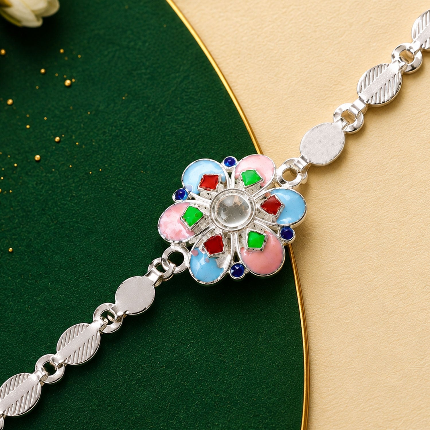 A silver chain rakhi with a colorful enamel flower charm on a dark green and gold background.
