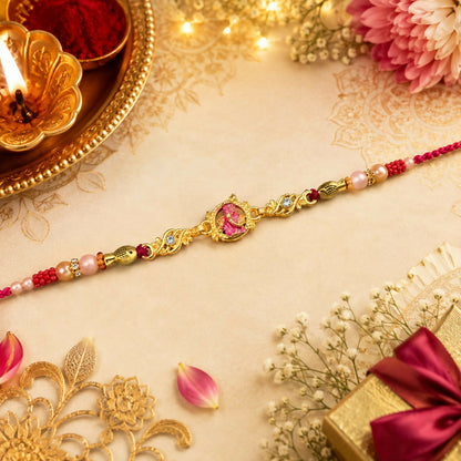 A gold and pink Lord Krishna Rakhi with red and white beads is centered on a light patterned background with flowers, rose
