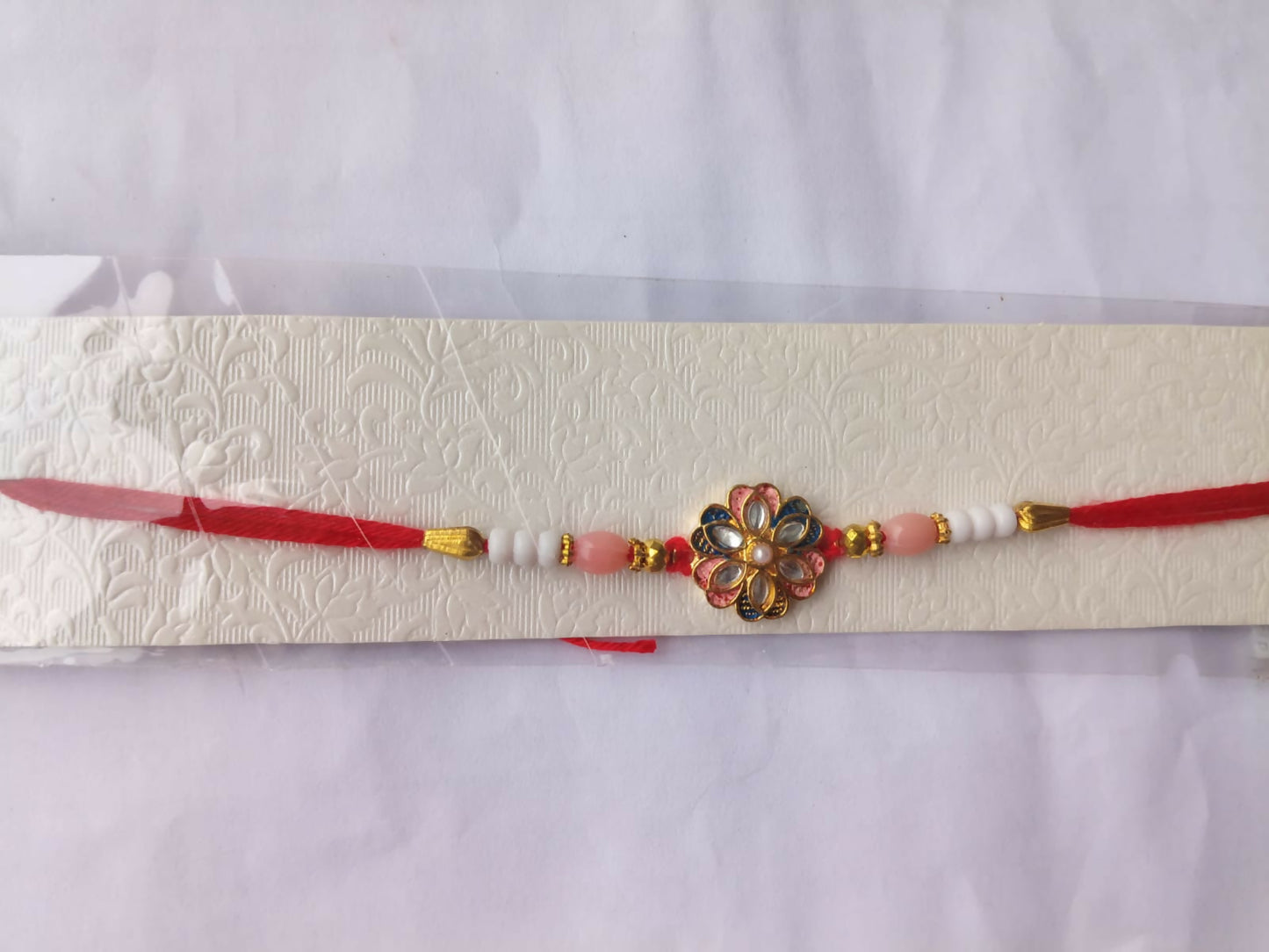A red thread with pink and white beads, gold accents, and a central floral charm with pink, blue, and clear stones, is