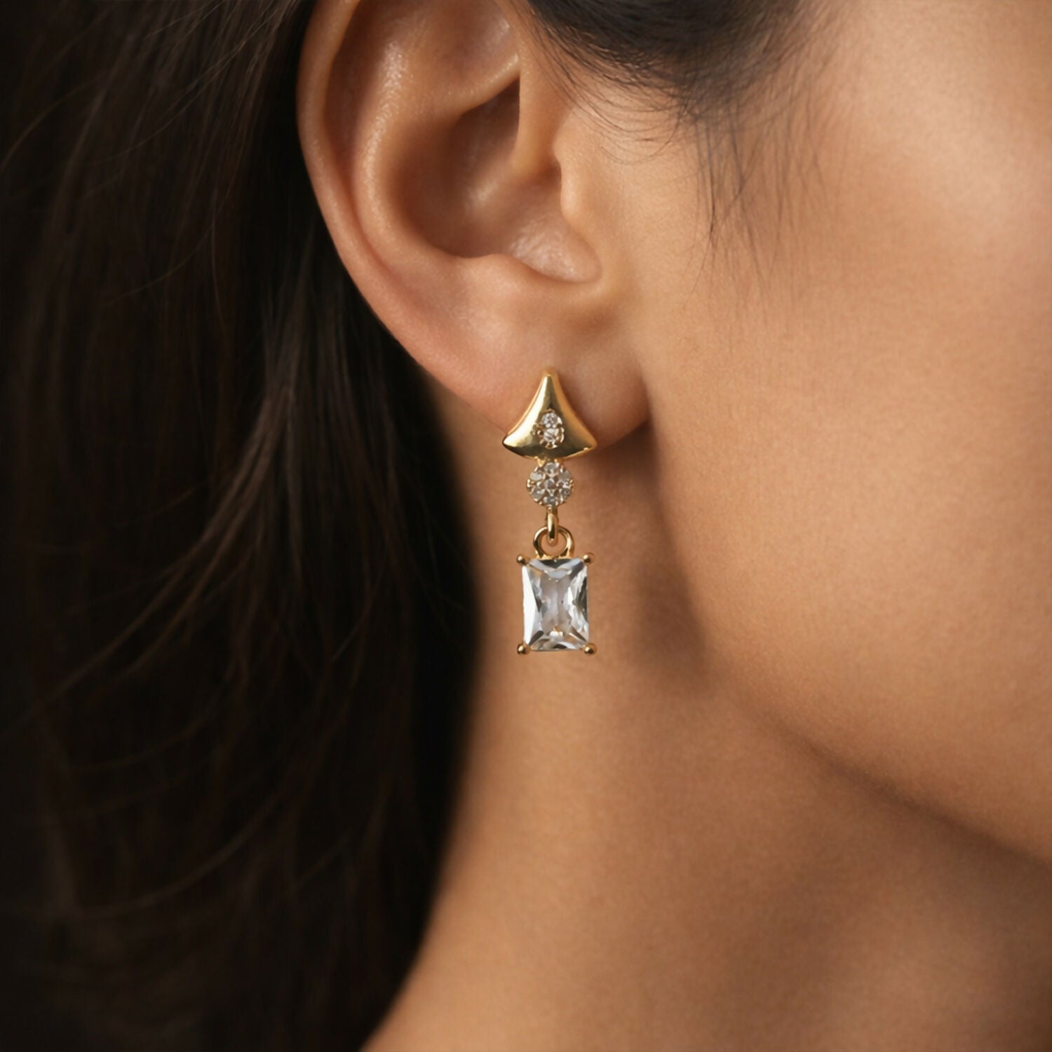Close-up of gold tone geometric crystal earrings with clear rectangular stone drop