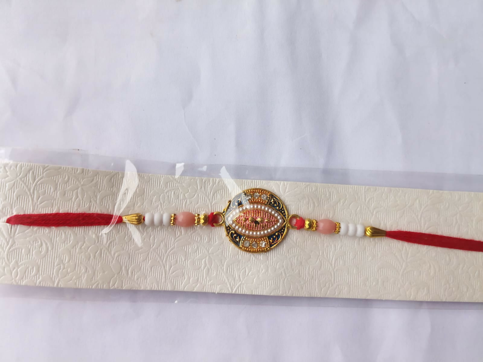 A blue, pink, and gold Rakhi with a central oval design, surrounded by small white stones and pearls, is displayed on a