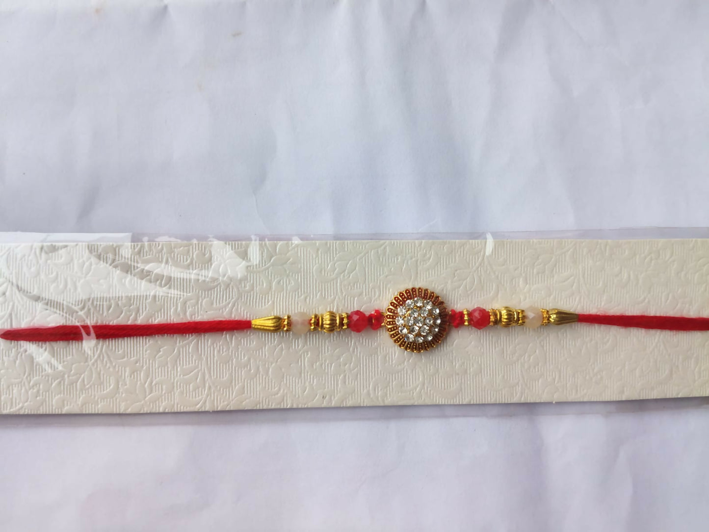 A red and gold bracelet with a central crystal medallion is presented on a textured white card against a plain background.