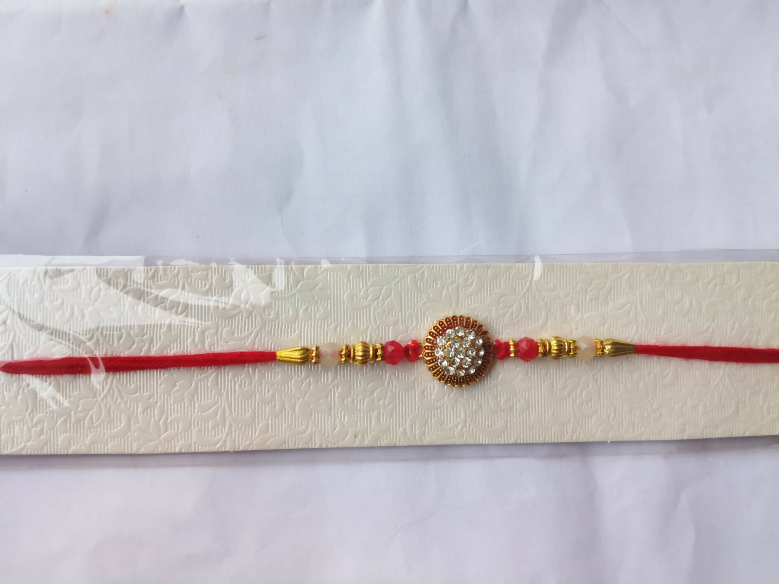 A red and gold bracelet with a central crystal medallion is presented on a textured white card against a plain background.