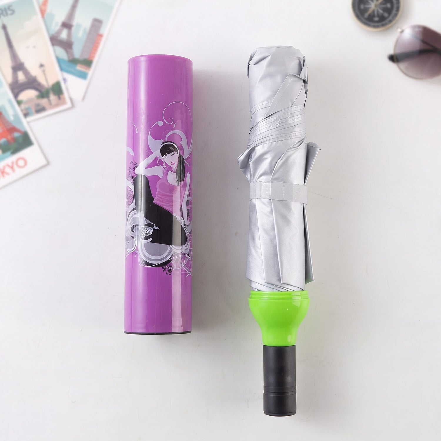 Collapsed grey umbrella with green handle next to its purple bottle-shaped case with a woman illustration, on a white