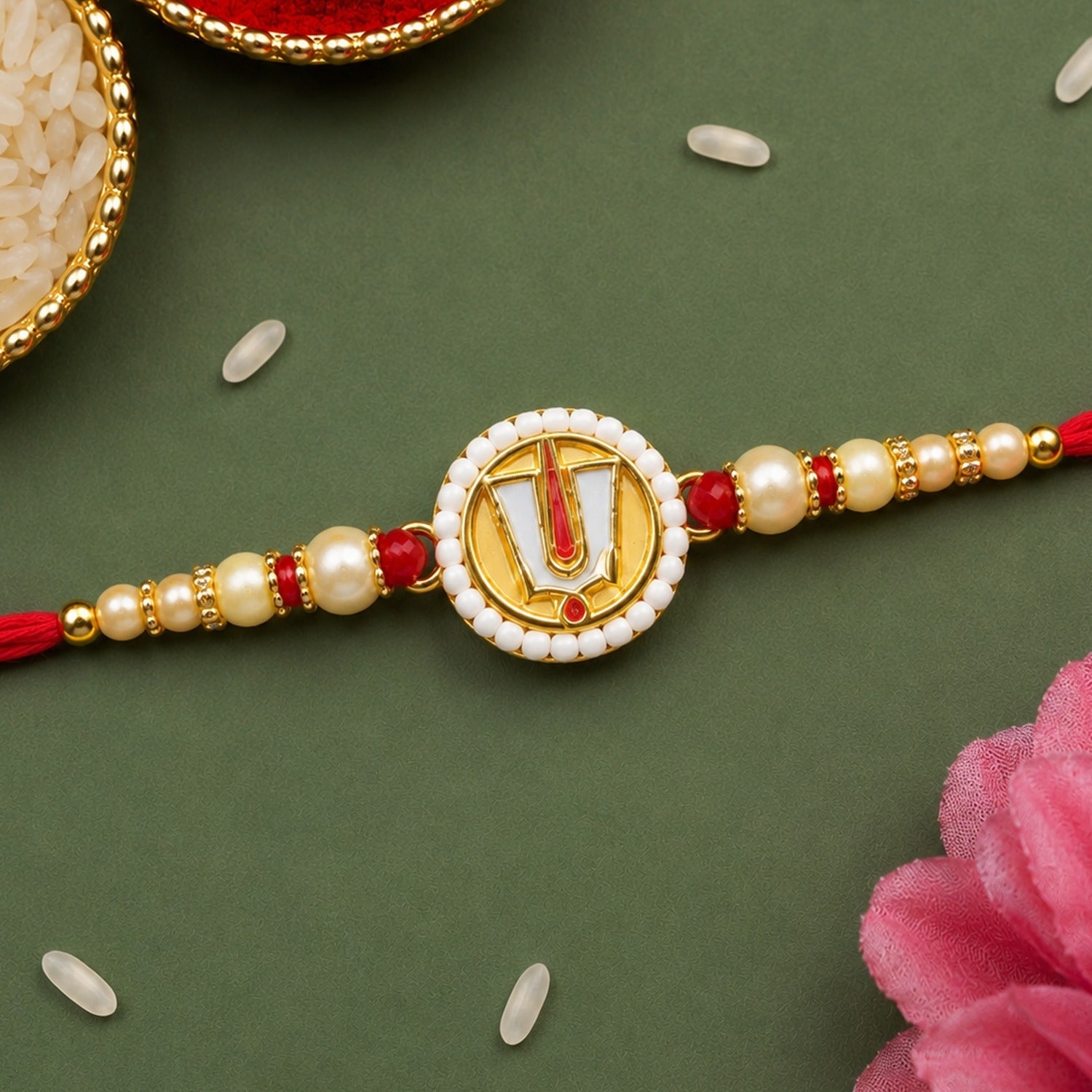 A single Rakhi with a red string, gold beads, and a circular charm on a dark green surface with scattered rice grains.