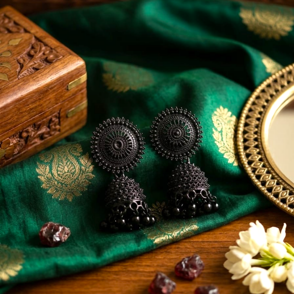 Two black oxidised jhumka earrings rest on a rich green and gold patterned cloth, surrounded by various trinkets.