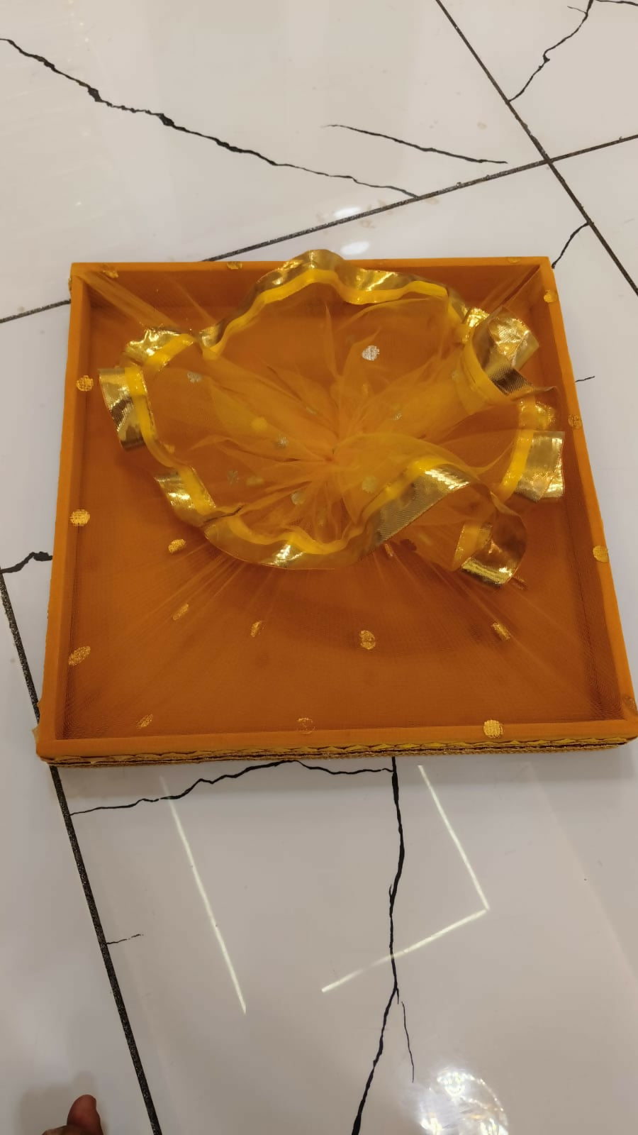 A yellow square tray with ruffled net and gold border, on a white tiled floor.