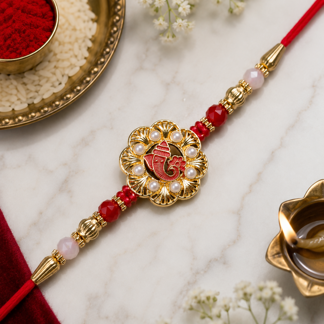 A red and gold Ganesha bracelet with red beads and golden detailing rests on a white marble surface, with a lit oil lamp in