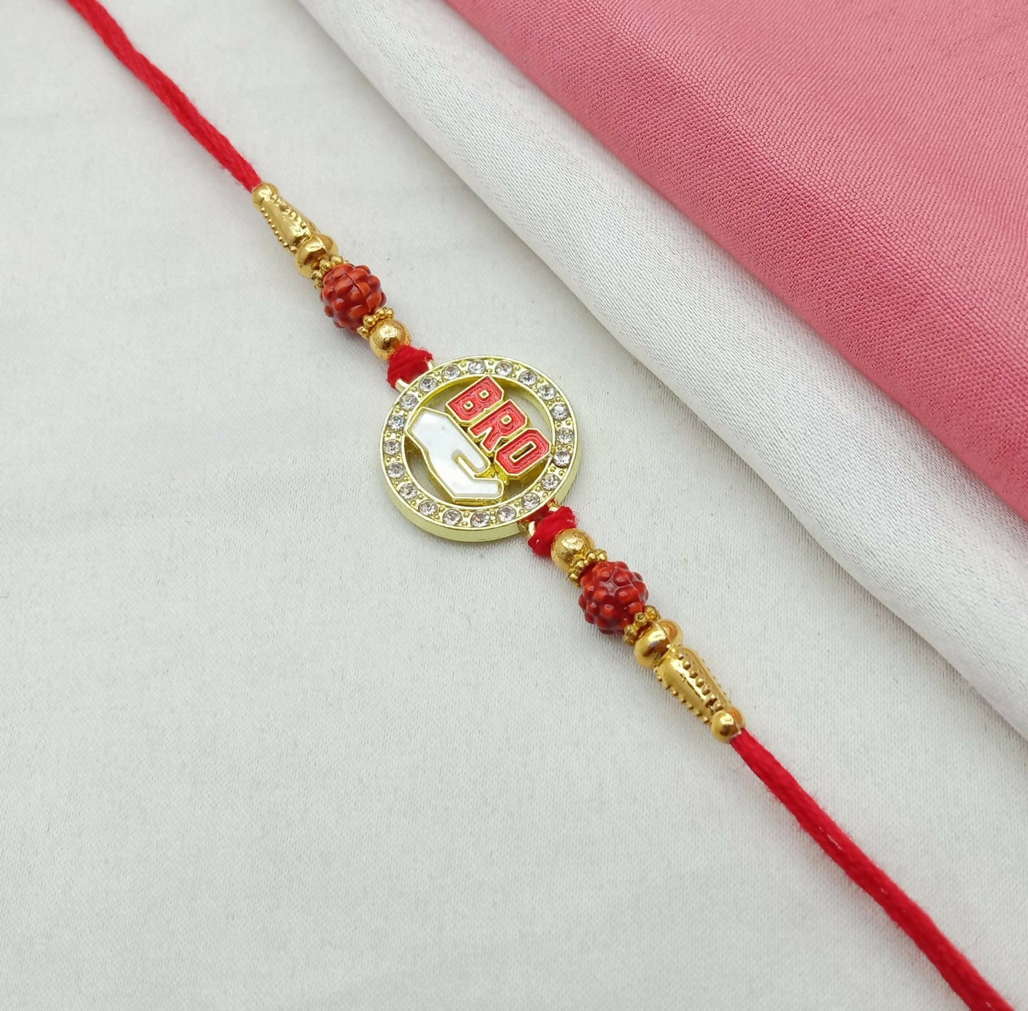 A red thread bracelet with a golden circular "BRO" charm, adorned with rhinestones and red carved beads, on a white and pink