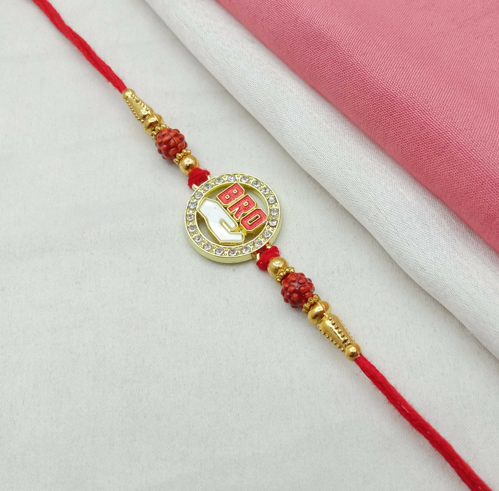 A red thread bracelet with a golden circular "BRO" charm, adorned with rhinestones and red carved beads, on a white and pink
