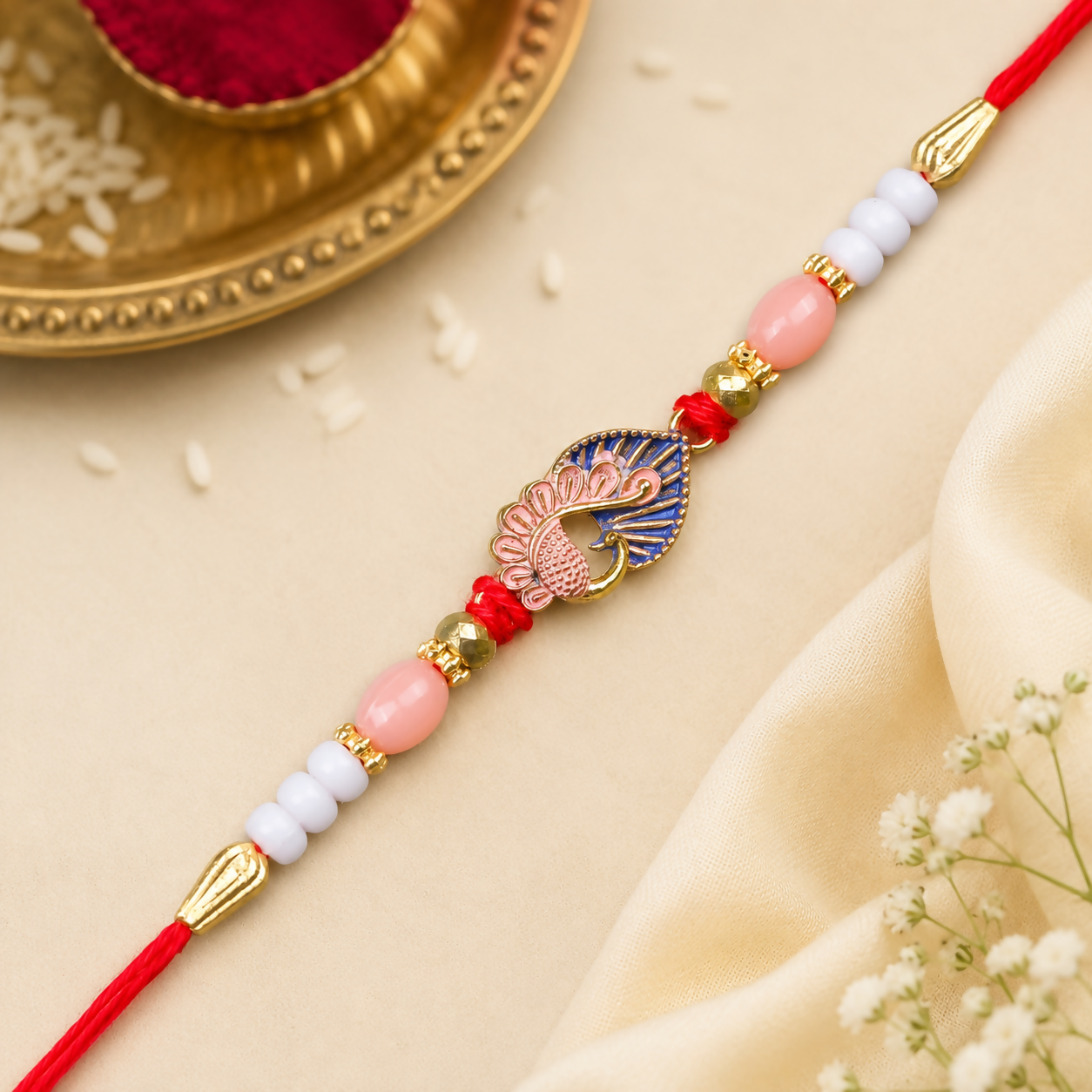 A red thread with a gold and blue peacock charm, flanked by pink, white, and gold beads, rests on a cream fabric.