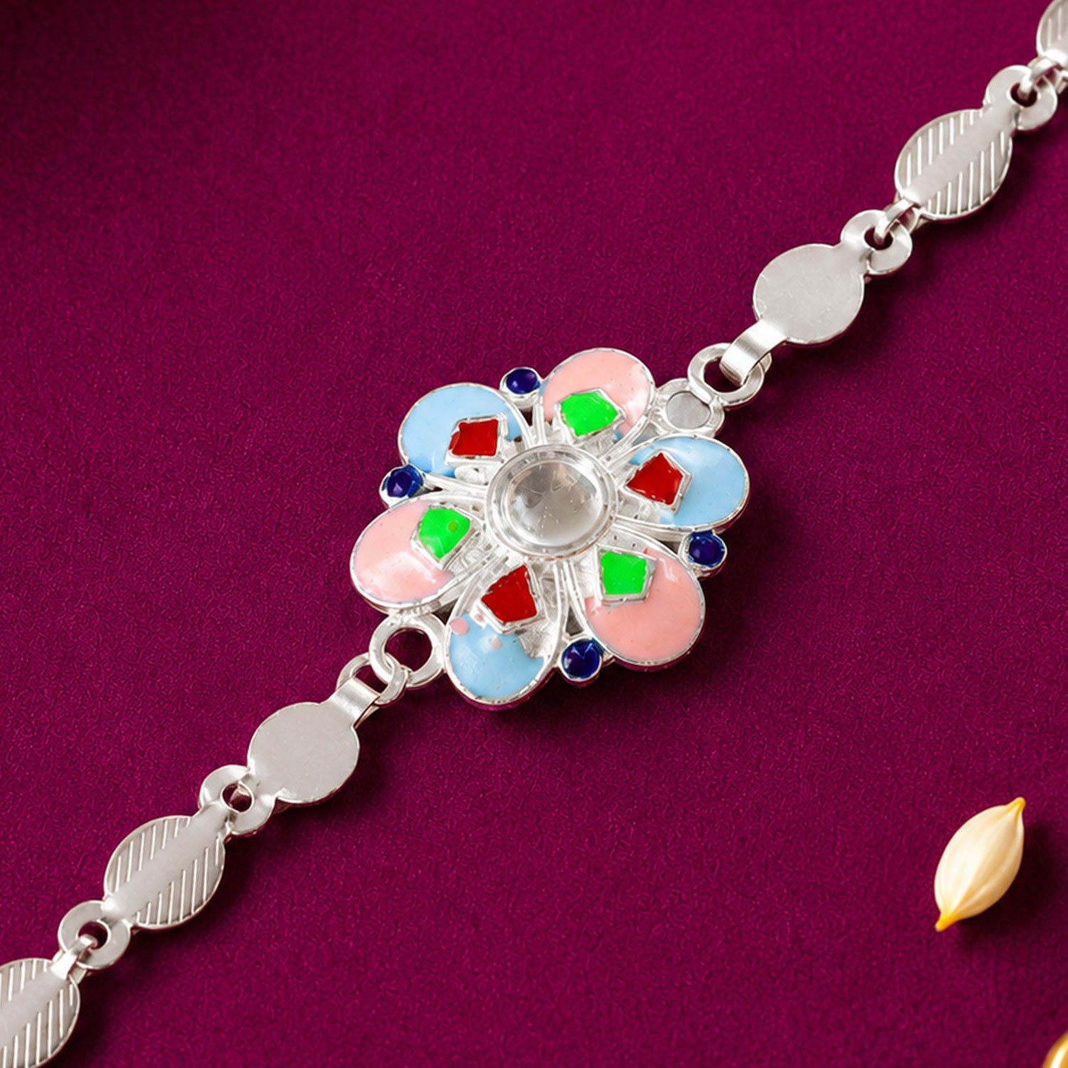 A silver chain rakhi with a colorful enamel flower charm on a deep purple background with a rice grain.