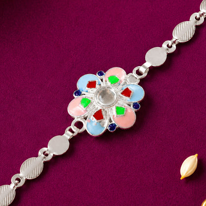 A silver chain rakhi with a colorful enamel flower charm on a deep purple background with a rice grain.