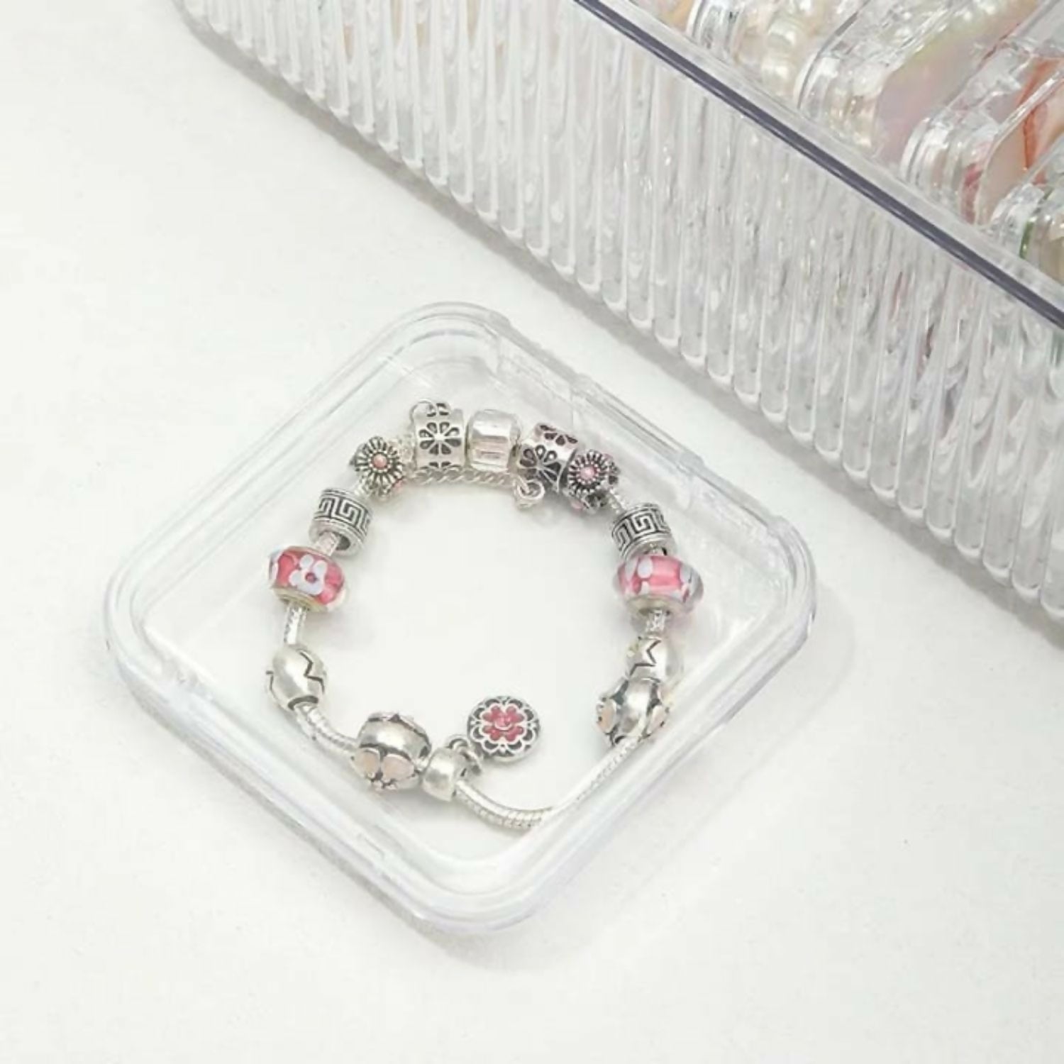 A silver charm bracelet with pink and white beads rests inside a small, clear square container.