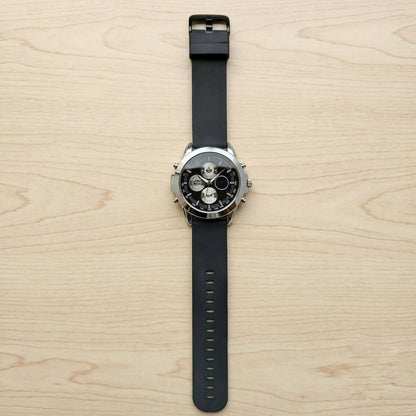 Top-down view of a black silicone strap watch with a silver case and black dial, on a light wood surface.