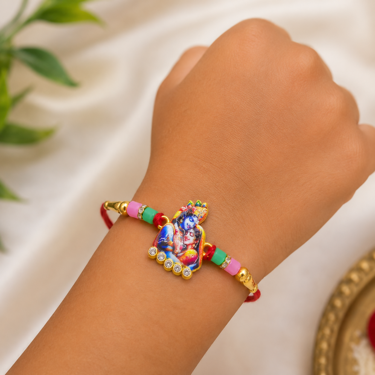 A child's wrist wearing a red thread with a golden Radha Krishna charm, on a white silk background with green leaves.