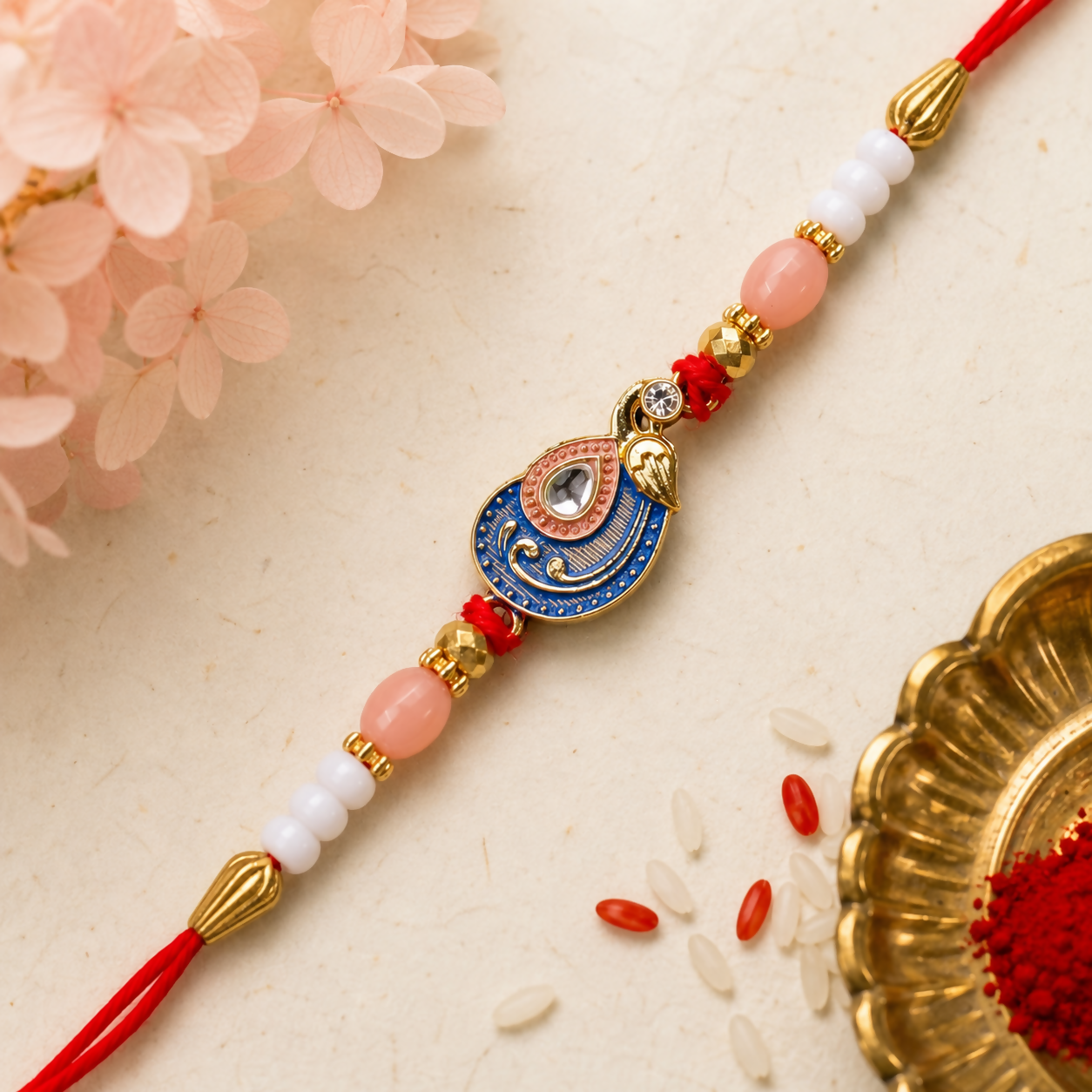 A red, blue, and gold peacock charm bracelet with pink and white beads is displayed on a textured beige surface with pink