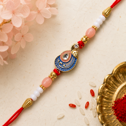 A red, blue, and gold peacock charm bracelet with pink and white beads is displayed on a textured beige surface with pink