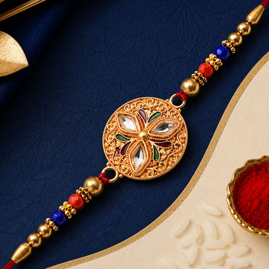 A gold-toned Meenakari rakhi with clear stones and colorful enamel, surrounded by red and blue beads on a red thread,