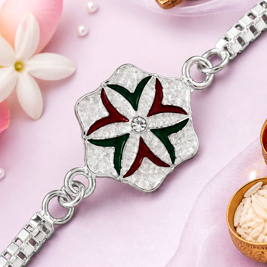 A silver rakhi with a flower design in red and green enamel, centered with a clear stone, on a pink background with white