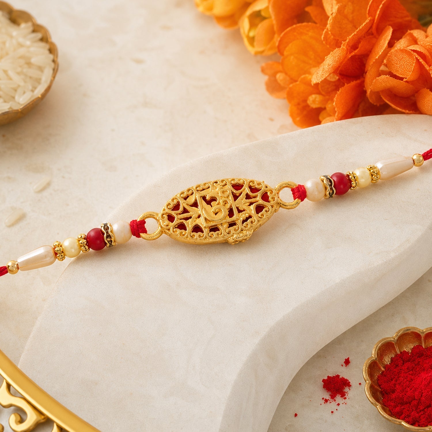 The gold Om charm with red filigree and beaded red and cream thread is displayed on a white marble surface with rice and red