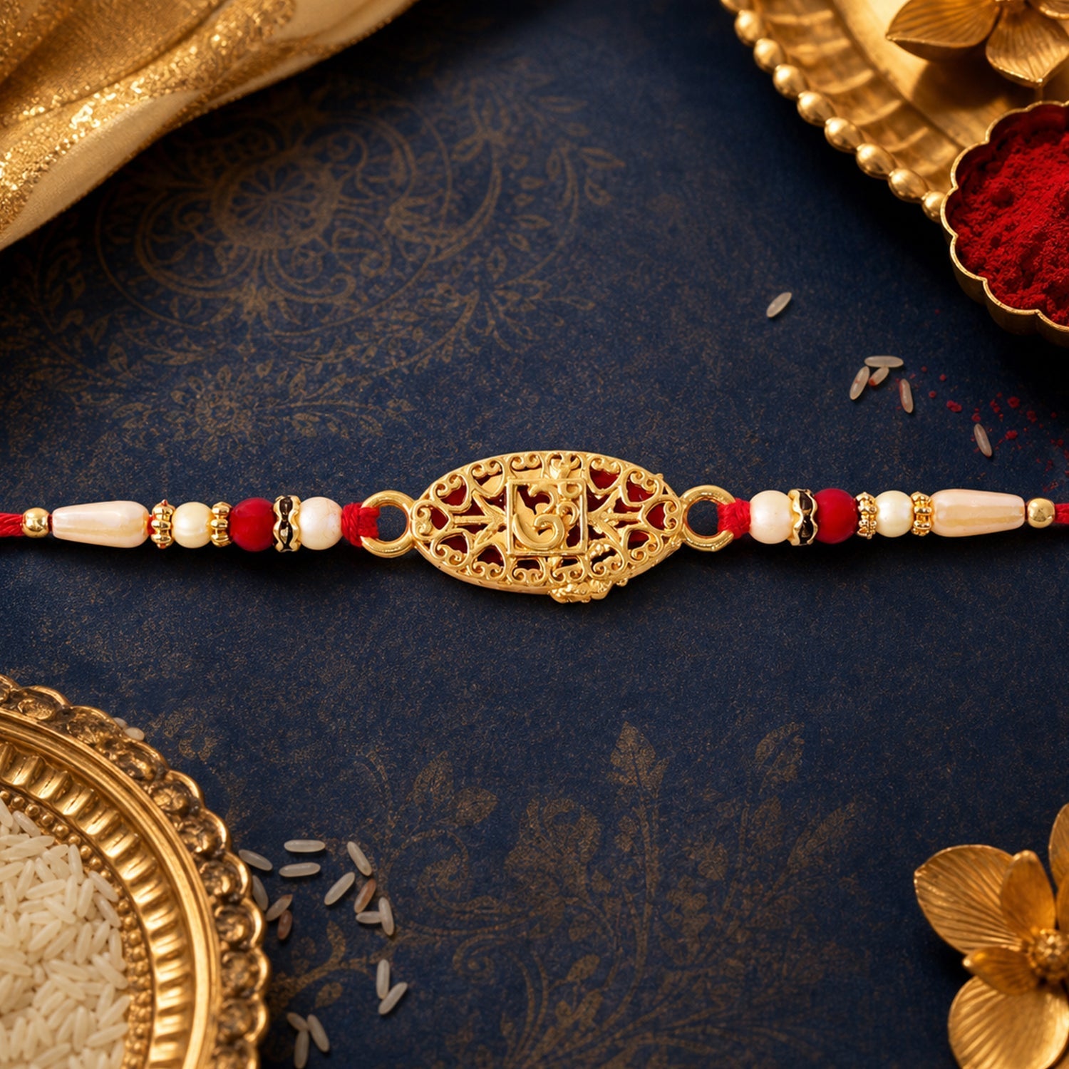 A gold Om charm with red filigree, flanked by red and cream beads on a red thread, rests on a blue patterned surface with