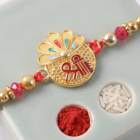A gold and red Rakhi with a central Shri symbol, red crystals, and gold beads, resting on a grey surface with small bowls of