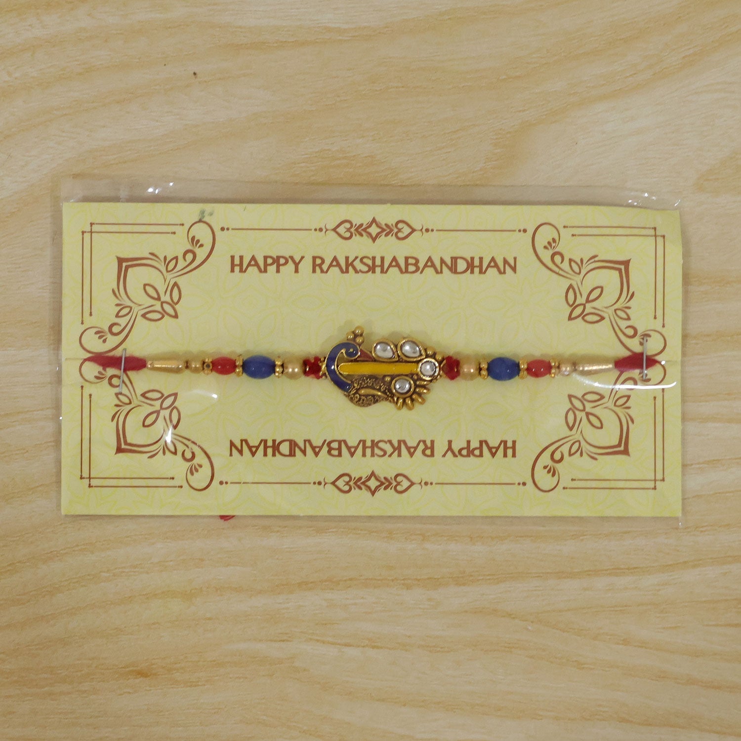 A red, blue, and gold beaded bracelet with a peacock charm is packaged on a yellow