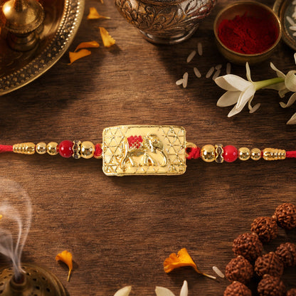 A gold elephant motif on a textured rectangular charm, flanked by gold and red beads on a red thread, rests on a wooden