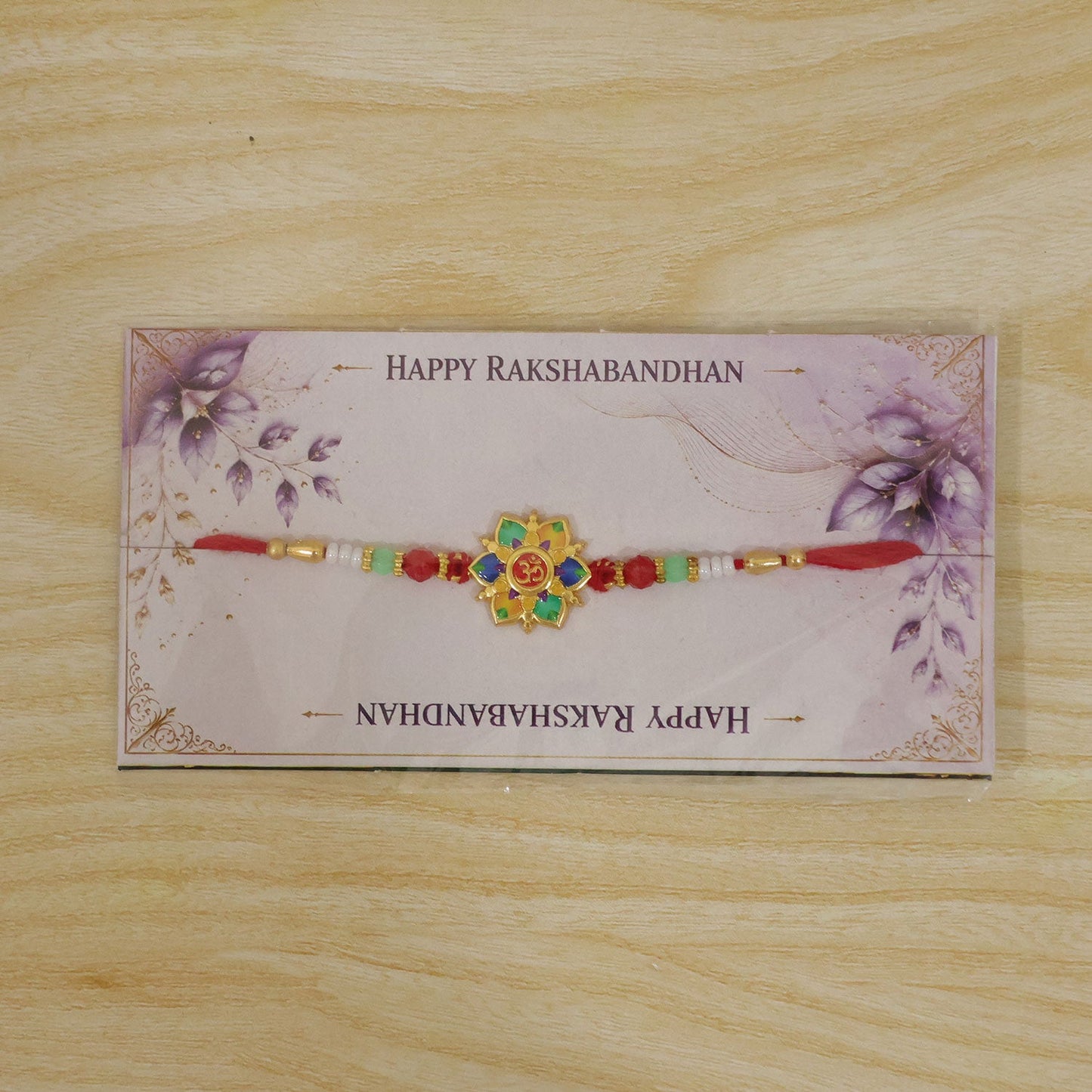 The colorful lotus and Om design, with red, green, and white beads on a red thread, is packaged on a decorative card with