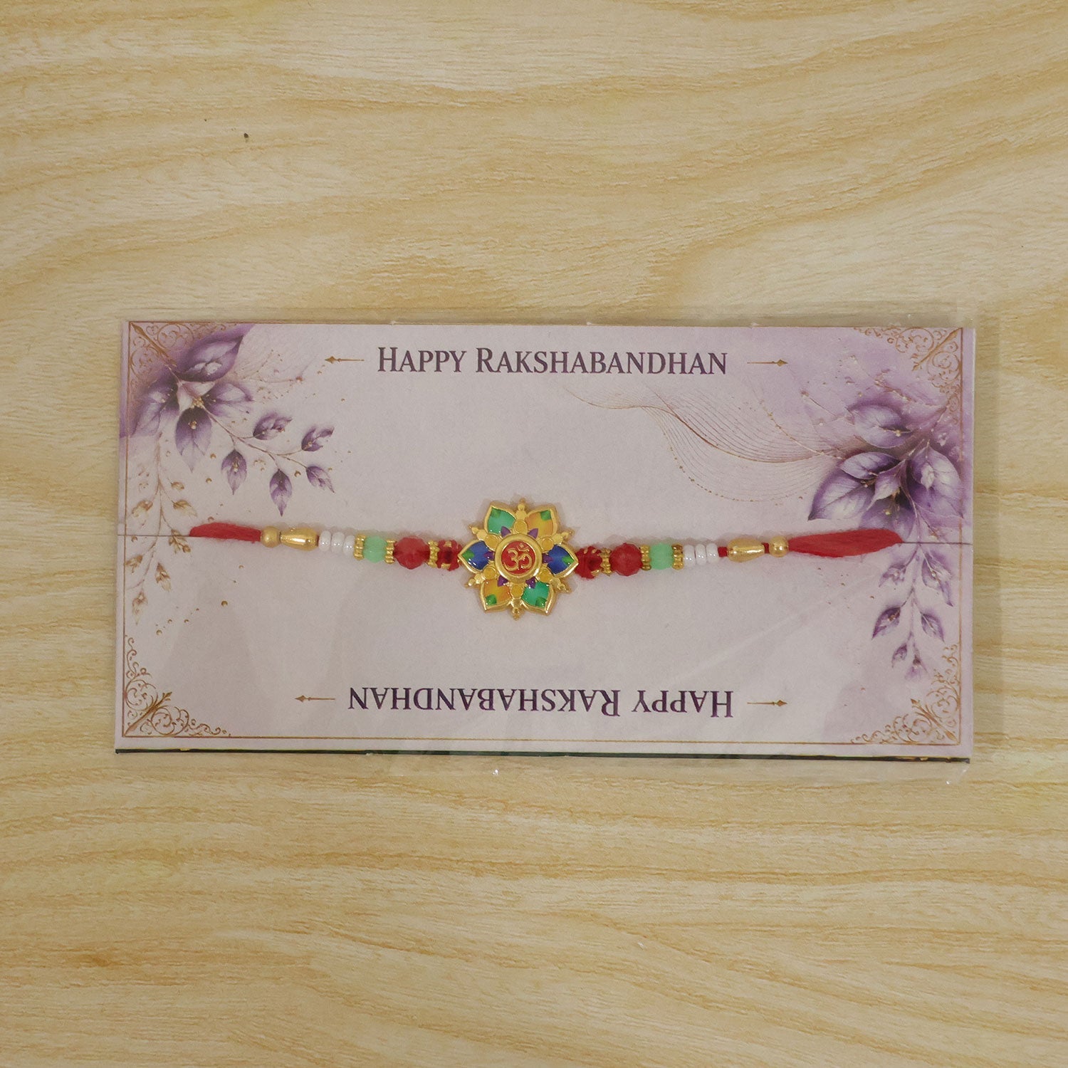 The colorful lotus and Om design, with red, green, and white beads on a red thread, is packaged on a decorative card with