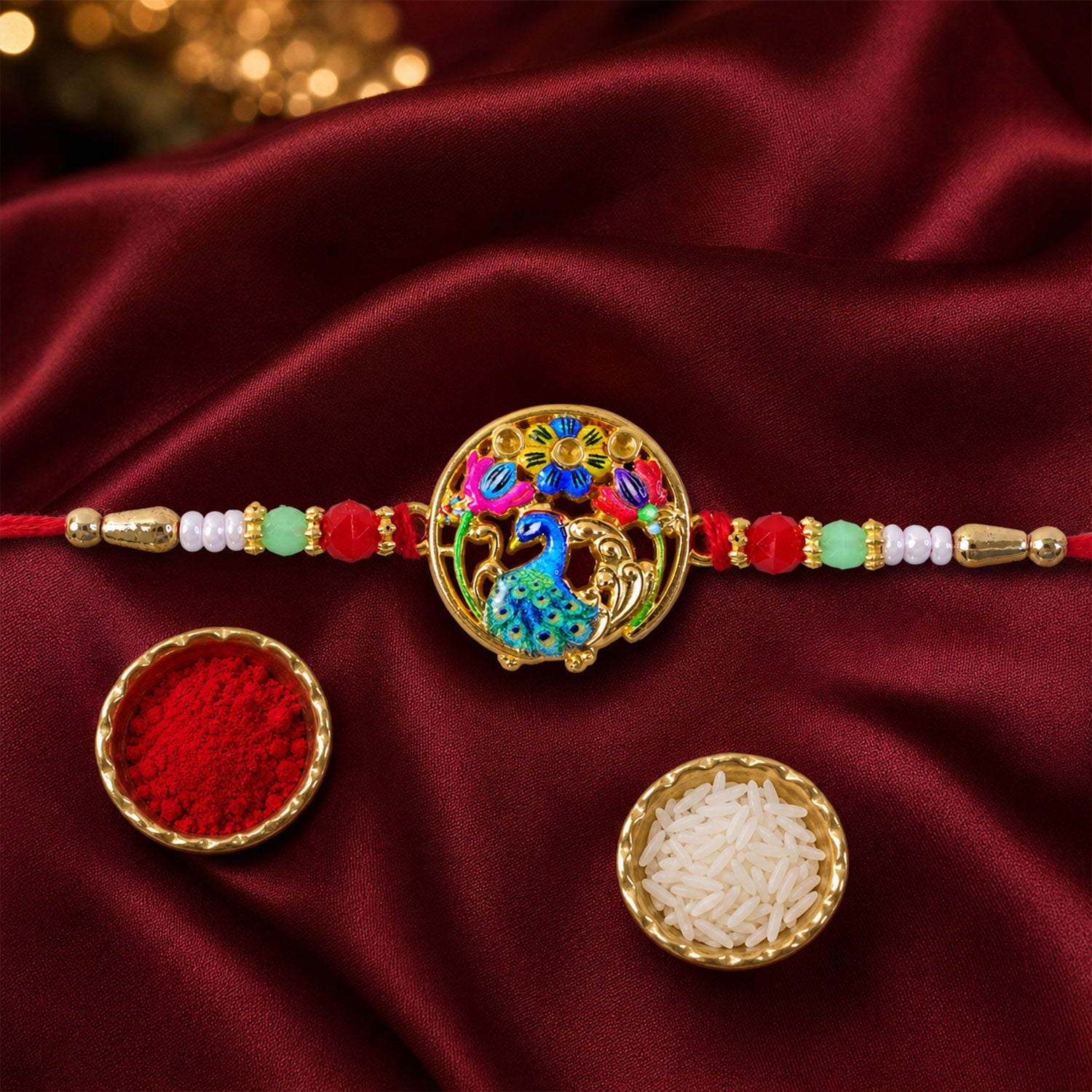 A gold-colored peacock and floral design on a red thread with colorful beads, next to small bowls of red powder and white