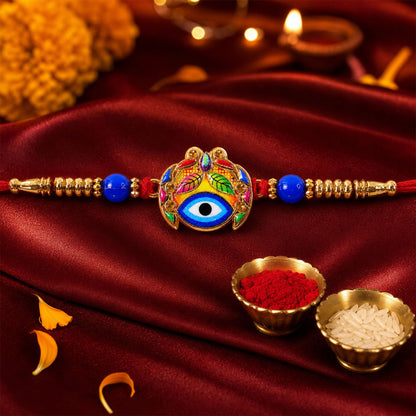 The Evil Eye Rakhi, featuring a blue eye and multicolored leaf design, is centered on a deep red satin cloth, with small