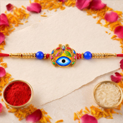 A colorful Evil Eye Rakhi with a prominent blue eye and leaf design, flanked by blue beads and gold elements, lies on a