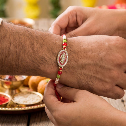A person's hand ties a red, gold, and pearl beaded bracelet with an oval centerpiece onto another person's wrist.