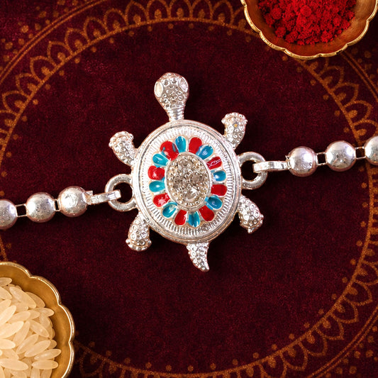 A silver-colored tortoise motif Rakhi with red and blue enamel details, surrounded by small bowls of rice and red powder on