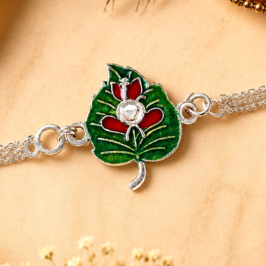 A silver chain bracelet features a green enamel leaf charm with a red Ganesha motif and a clear stone center.