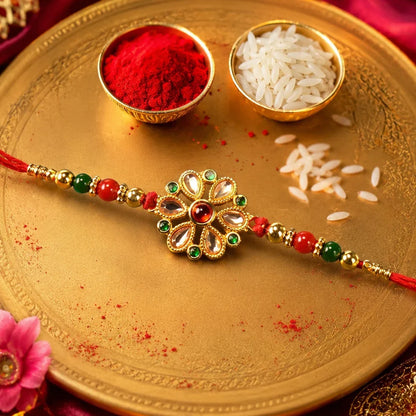 A red bracelet with a floral Kundan charm and red and green beads lies on a golden tray with rice and red powder.