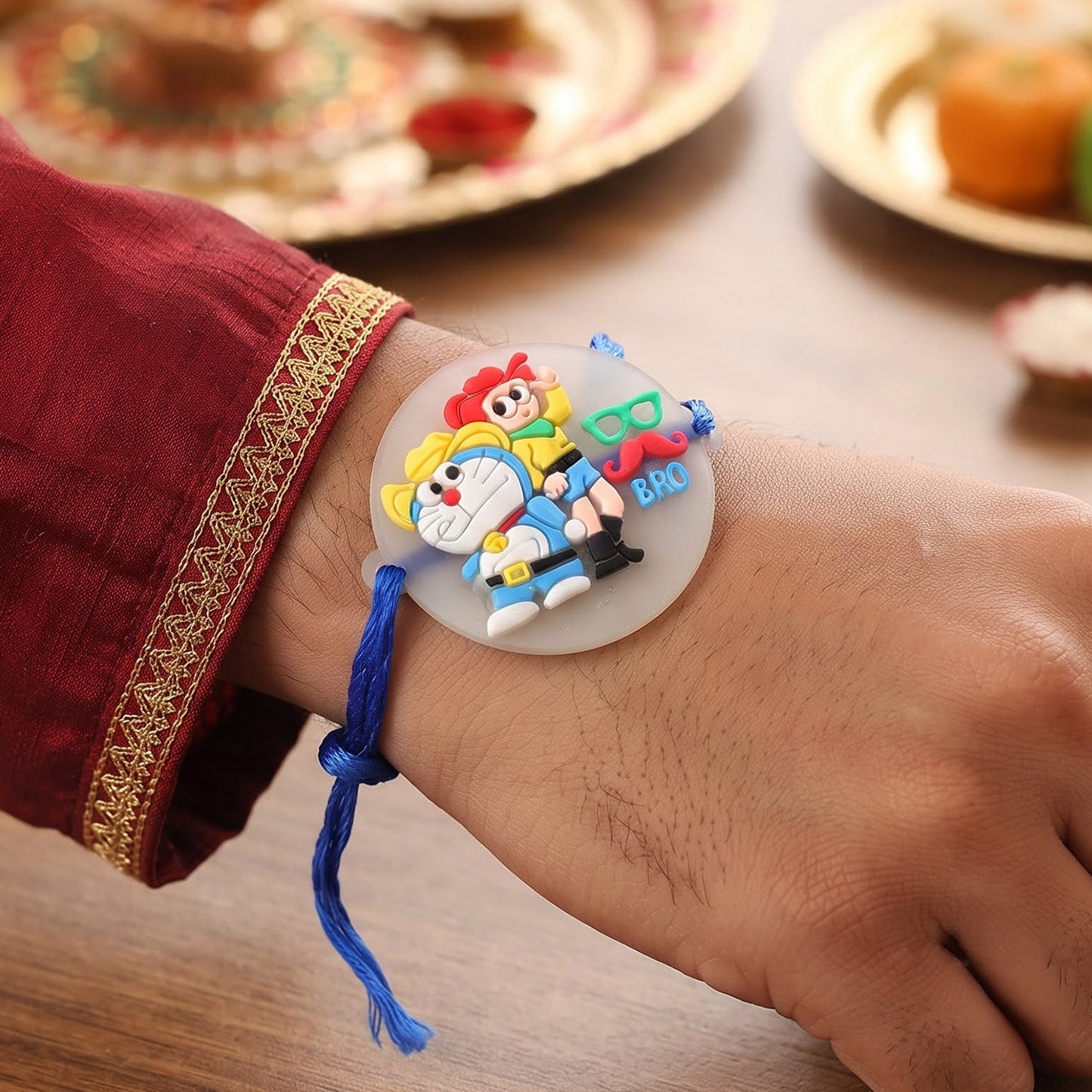 A person's wrist wearing the blue Doraemon and Nobita Rakhi with a 'BRO' charm, against a blurred background.
