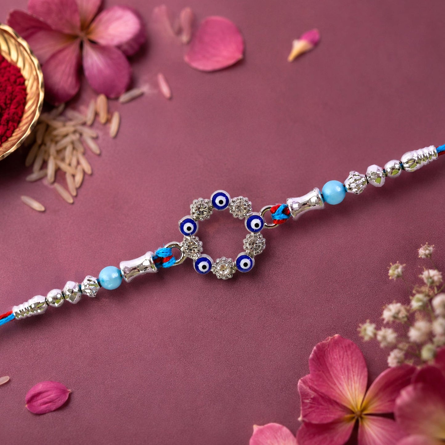 A blue and red thread rakhi with a circular evil eye charm, silver and turquoise beads on a dark pink floral background.