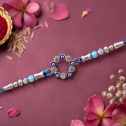 A blue and red thread rakhi with a circular evil eye charm, silver and turquoise beads on a dark pink floral background.