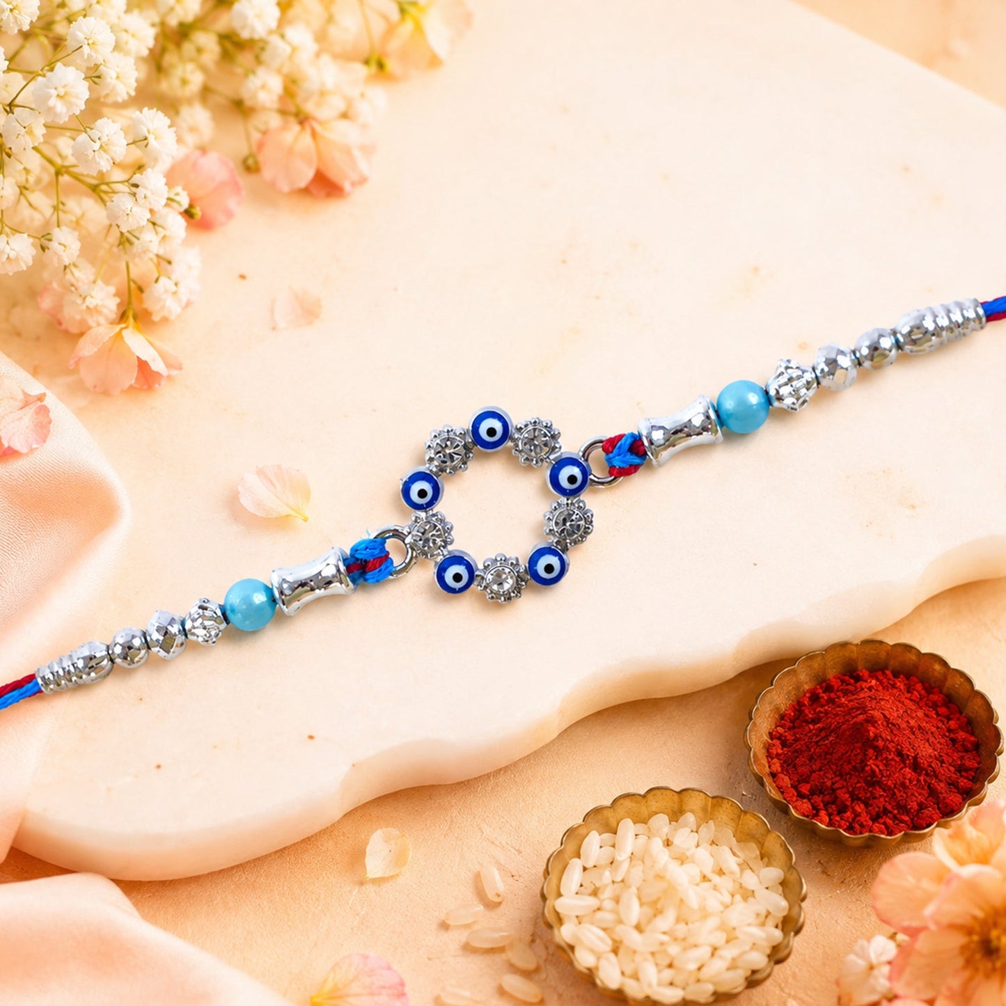 A blue and red thread rakhi with a circular evil eye charm, silver and turquoise beads on a light floral background with
