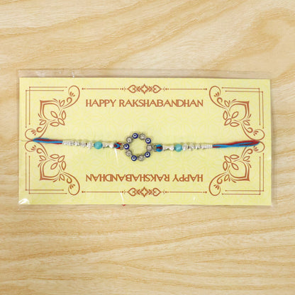 A blue and red thread rakhi with a circular evil eye charm, silver and turquoise beads on a yellow Happy Rakshabandhan card.